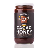 Front view of a 1-pound jar of Sauce Bae Cacao Honey, featuring a cartoon chocolate character on a black label with the text “Cacao Honey – Raw Honey + Cacao + Vanilla.